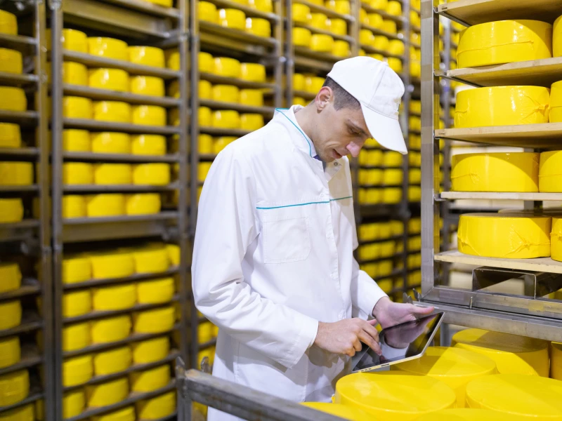Technoloog in witte jas die naast kasten met kazen staat en daar uitdagingen noteert in de sector Food & Pharma